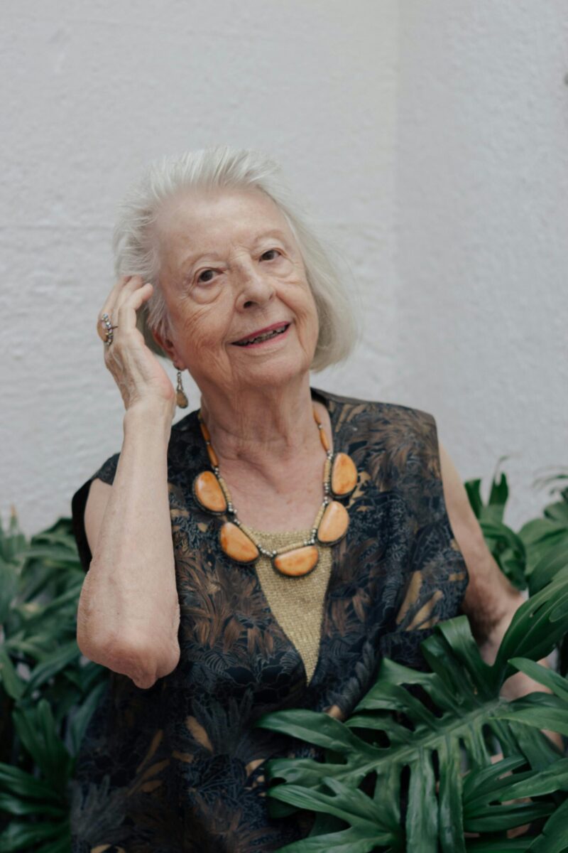 Portrait of a stylish elderly woman wearing a statement necklace, surrounded by greenery.